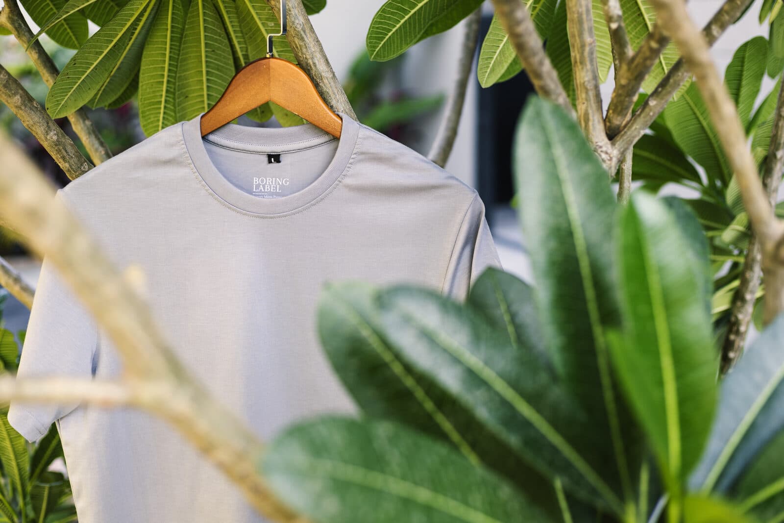 Boring Label 100% Supima cotton round neck t-shirt in light grey, hanging on a wooden hanger among lush green plants