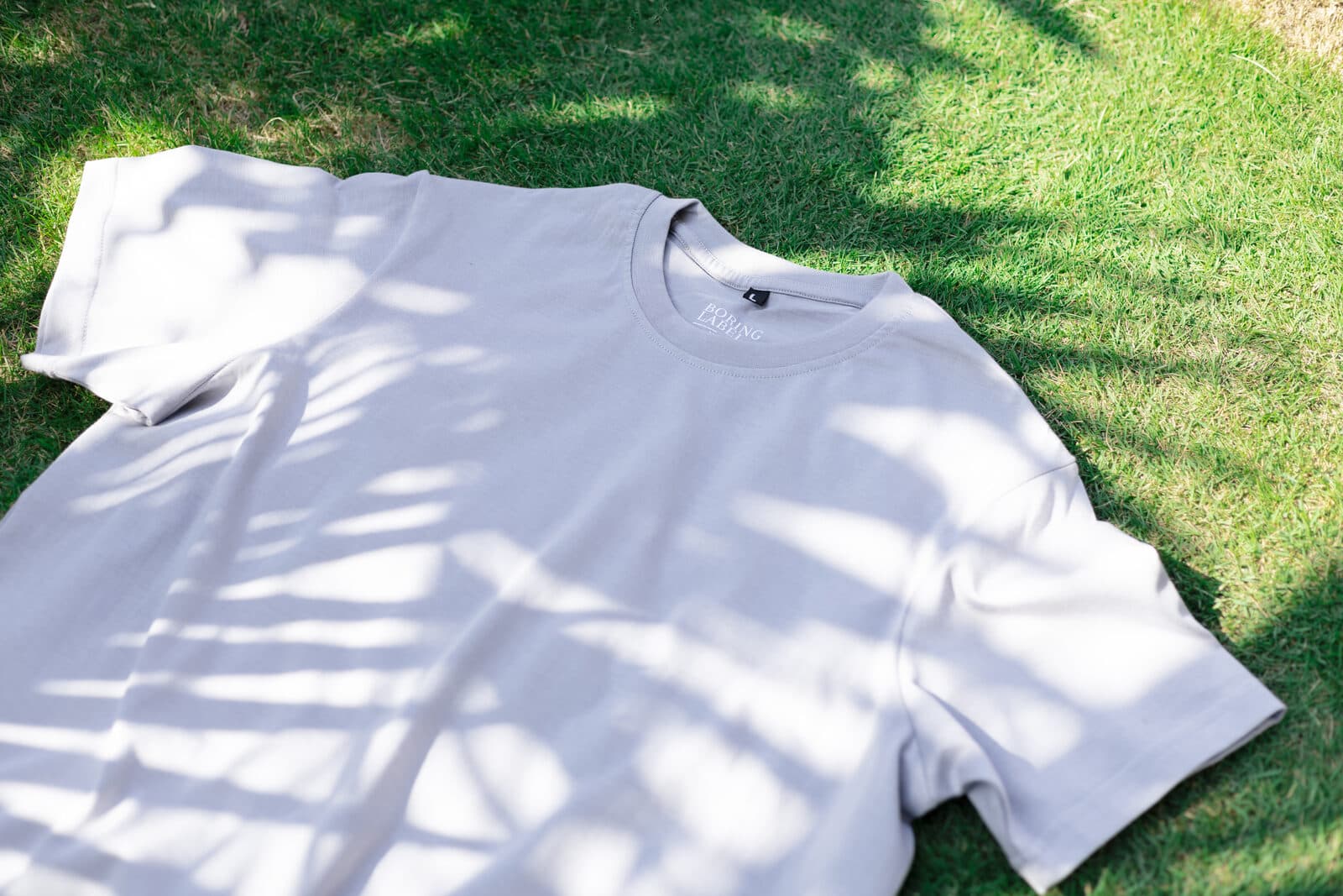 Boring Label premium Supima cotton t-shirt laid flat on sunlit grass with natural tree shadow patterns