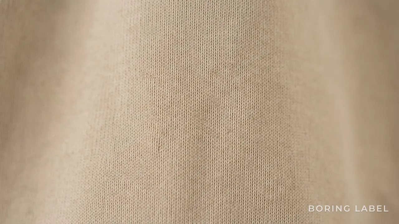 Close-up macro of densely knit plain cotton jersey fabric showing a smooth durable surface, neutral sand tone, soft diffused daylight, no markings