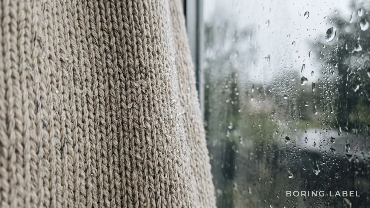Close-up of light cotton knit fabric texture beside a rain-misted window, soft grey daylight, droplets of water on the weave, neutral oatmeal palette, calm editorial macro photography