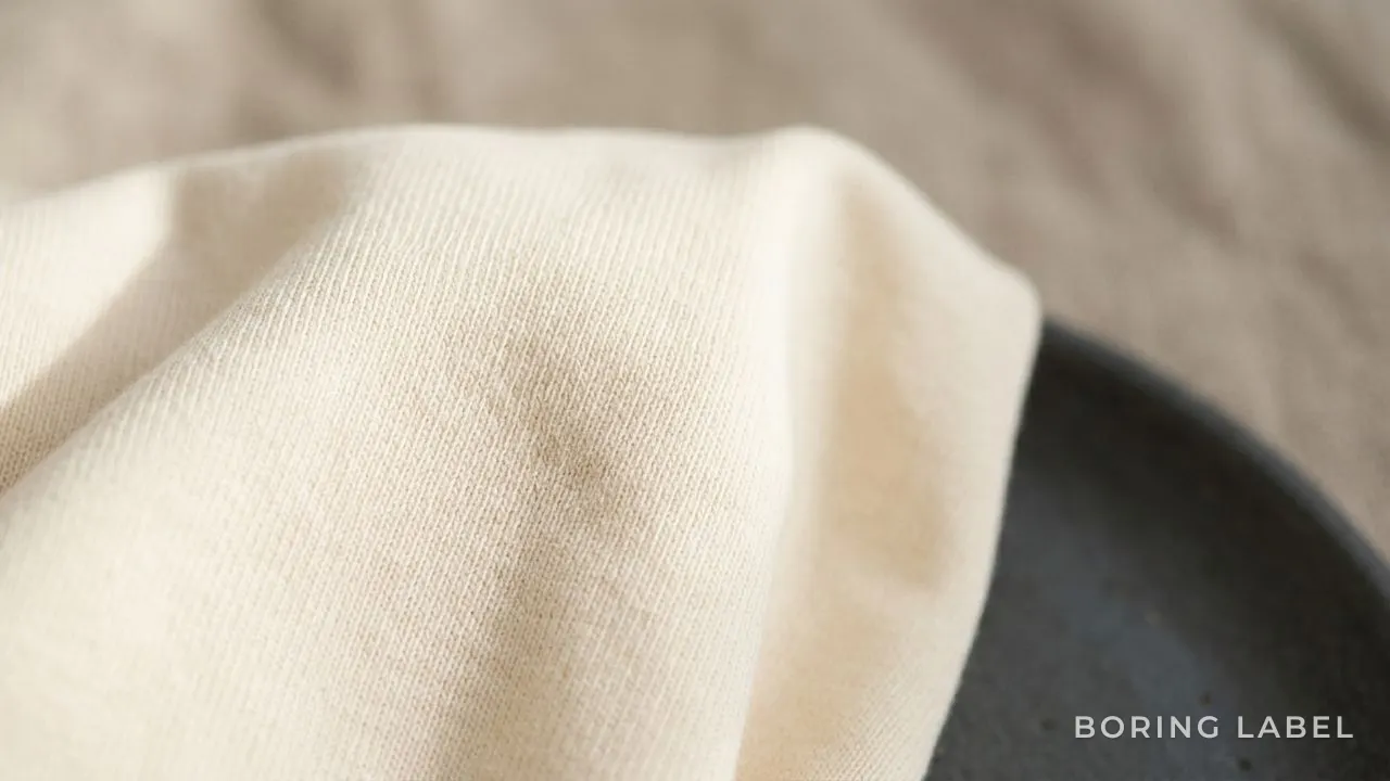 Macro detail of smooth long-staple cotton jersey fabric in soft cream, raking side light revealing a clean even surface with no fuzz, minimalist neutral palette, matte and premium